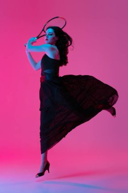 Siyah gece elbisesi giyen, tenis oynayan, pembe neon arka planda soyutlanmış, şık bir kadın. Moda ve spor anlayışı