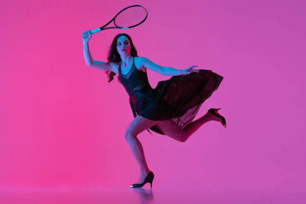 Alta moda estilo jovem mulher em vestido de noite preto, vestido jogando tênis isolado sobre fundo de néon rosa. Conceito de moda e esporte — Fotografia de Stock