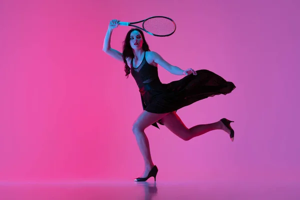 Hög mode stylad ung kvinna i svart aftonklänning, klänning spelar tennis isolerad över rosa neon bakgrund. Begreppet mode och idrott — Stockfoto