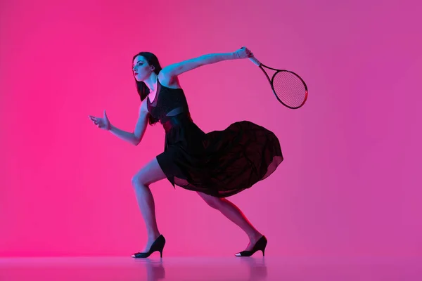 High-fashion styled young woman in black evening gown, dress playing tennis isolated over pink neon background. Concept of fashion and sport — Stock Photo, Image