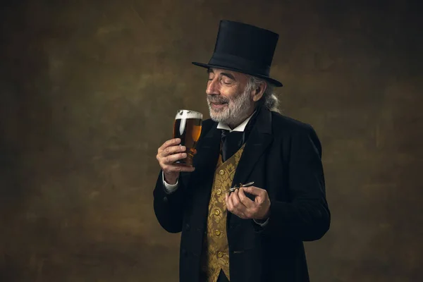 Uomo anziano dai capelli grigi, gentiluomo, aristocratico o attore che beve birra isolata su sfondo vintage scuro. Stile retrò, confronto del concetto di epoche. — Foto Stock