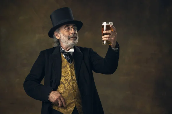 Homme âgé aux cheveux gris, gentleman, aristocrate ou acteur buvant de la bière isolée sur fond vintage sombre. Style rétro, comparaison des époques concept. — Photo