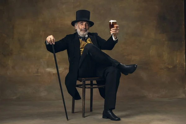 Älterer grauhaariger Mann, Gentleman, Aristokrat oder Schauspieler, der isoliert auf dunklem Vintage-Hintergrund Bier trinkt. Retro-Stil, Vergleich des Epochenkonzepts. — Stockfoto