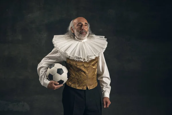 Älterer grauhaariger Mann, Schauspieler, der isoliert mit Fußballball auf dunklem Vintage-Hintergrund posiert. Retro-Stil, Vergleich des Epochenkonzepts. — Stockfoto