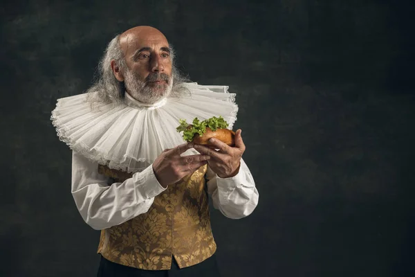 Um homem idoso de cabelos grisalhos, pessoa histórica medieval, ator degustando fast food isolado em fundo escuro vintage. Estilo retro, comparação do conceito eras. — Fotografia de Stock