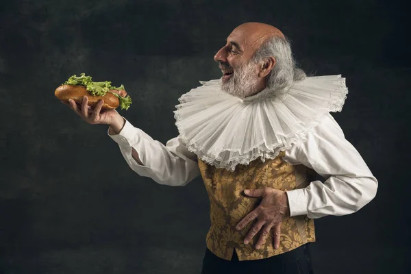 Um homem idoso de cabelos grisalhos, pessoa histórica medieval, ator degustando fast food isolado em fundo escuro vintage. Estilo retro, comparação do conceito eras. — Fotografia de Stock