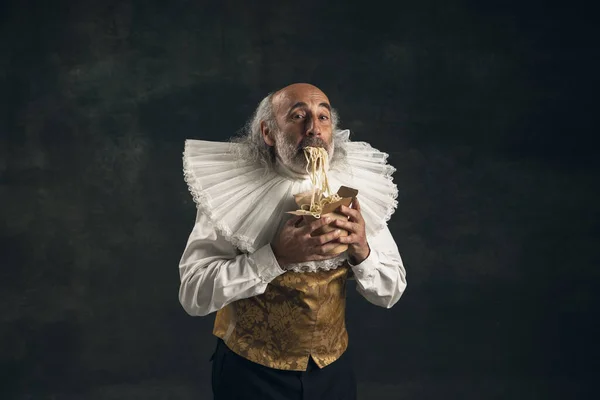 Um homem idoso de cabelos grisalhos, pessoa histórica medieval, ator degustando fast food isolado em fundo escuro vintage. Estilo retro, comparação do conceito eras. — Fotografia de Stock