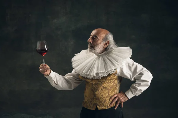 Homem idoso de cabelos grisalhos, pessoa histórica medieval, ator bebendo vinho isolado em fundo escuro vintage. Estilo retro, comparação do conceito eras. — Fotografia de Stock