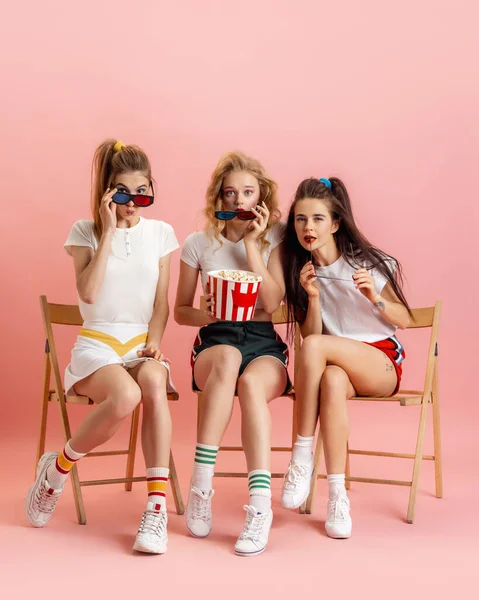 Três mulheres jovens em retro estilo de moda dos anos 90, roupas assistindo filme isolado sobre fundo de estúdio rosa. Conceito de comparação eras, beleza, moda e juventude. — Fotografia de Stock