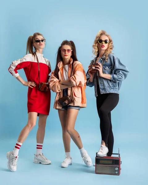 Hübsche junge Frauen im Retro-Stil der 90er Jahre, Outfits, die isoliert vor blauem Studiohintergrund posieren. Konzept des Epochenvergleichs, Schönheit, Mode und Jugend. — Stockfoto