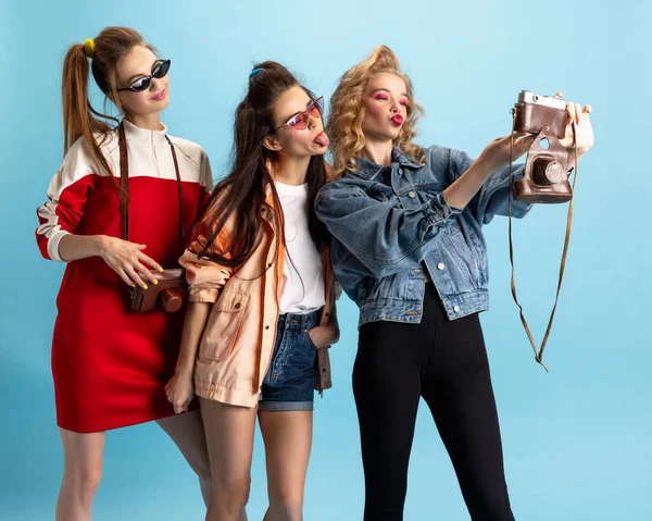 Drei junge Mädchen im Retro-Stil der 90er Jahre, Outfits, die isoliert vor blauem Studiohintergrund posieren. Konzept des Epochenvergleichs, Schönheit, Mode und Jugend. — Stockfoto