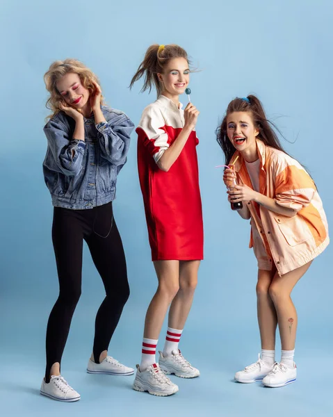 Mujeres bastante jóvenes en estilo de moda retro de los 90, trajes posando aislados sobre fondo de estudio azul. Concepto de eras comparación, belleza, moda y juventud. — Foto de Stock