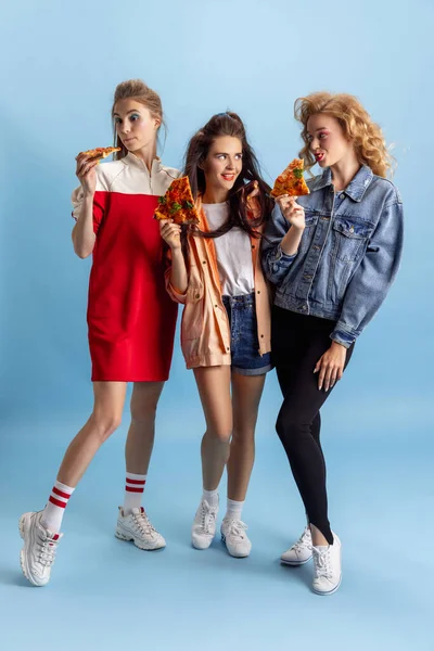 Tres mujeres atractivas jóvenes en estilo de moda retro de los 90, trajes comiendo pizza aislados sobre fondo azul del estudio. Concepto de eras comparación, belleza, moda y juventud. —  Fotos de Stock