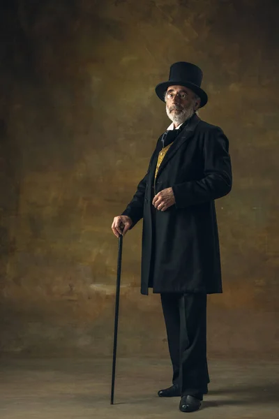Portrait of elderly gray-haired man, gentleman, aristocrat or actor posing isolated on dark vintage background. Retro style, comparison of eras concept.