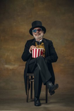 Cute senior smiling man, gentleman, aristocrat or actor watching movie isolated on dark vintage background. Retro style, comparison of eras concept.