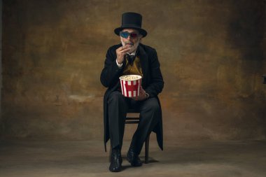 Cute senior smiling man, gentleman, aristocrat or actor watching movie isolated on dark vintage background. Retro style, comparison of eras concept.