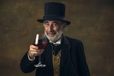 Portrait of elderly gray-haired man, gentleman, aristocrat or actor tasting wine isolated on dark vintage background. Retro style, comparison of eras concept.