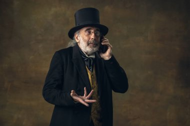 Cute senior man, gentleman, aristocrat or actor talking on phone isolated on dark vintage background. Retro style, comparison of eras concept.