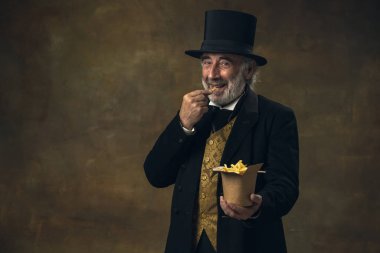 Handsome elderly gray-haired man, gentleman, aristocrat or actor eating fast food isolated on dark vintage background. Retro style, comparison of eras concept.