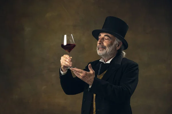 Portrait of elderly gray-haired man, gentleman, aristocrat or actor tasting wine isolated on dark vintage background. Retro style, comparison of eras concept. — Foto Stock