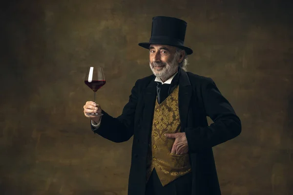 Portrait of elderly gray-haired man, gentleman, aristocrat or actor tasting wine isolated on dark vintage background. Retro style, comparison of eras concept. — Foto Stock