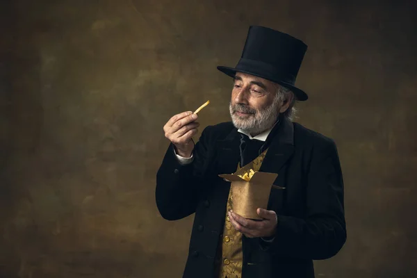 Handsome elderly gray-haired man, gentleman, aristocrat or actor eating fast food isolated on dark vintage background. Retro style, comparison of eras concept. — Zdjęcie stockowe