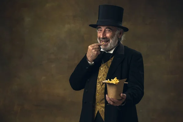 Handsome elderly gray-haired man, gentleman, aristocrat or actor eating fast food isolated on dark vintage background. Retro style, comparison of eras concept. — Foto Stock