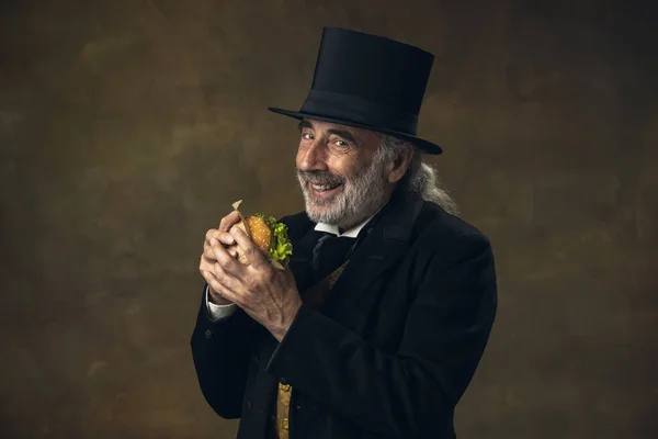 Handsome elderly gray-haired man, gentleman, aristocrat or actor eating fast food isolated on dark vintage background. Retro style, comparison of eras concept. — Foto Stock