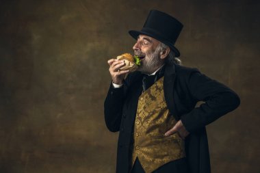 Handsome elderly gray-haired man, gentleman, aristocrat or actor eating fast food isolated on dark vintage background. Retro style, comparison of eras concept.
