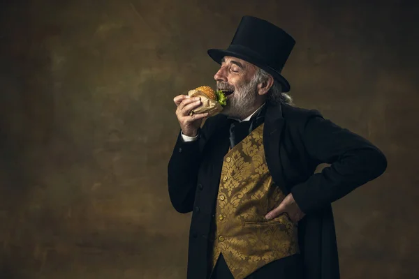 Handsome elderly gray-haired man, gentleman, aristocrat or actor eating fast food isolated on dark vintage background. Retro style, comparison of eras concept. — Foto de Stock