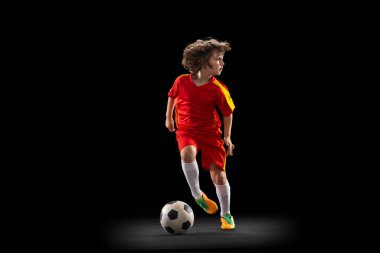 In motion. One little male football soccer player, boy training with football ball isolated on dark studio background. Concept of sport, game, hobby