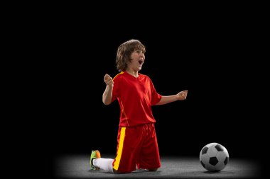 Happy, excited boy, football soccer player in red sports uniform with football ball isolated on dark studio background. Concept of sport, game, hobby