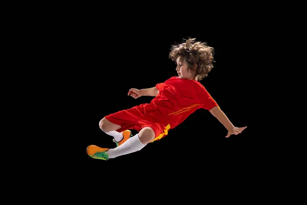 Preschool boy, football soccer player in red sports uniform practicing with football ball isolated on dark studio background. Concept of sport, game, hobby — Foto de Stock