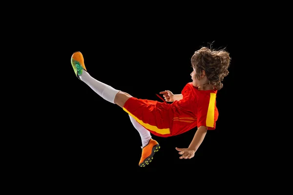 Menino pré-escolar, jogador de futebol em uniforme esportivo vermelho praticando com bola de futebol isolada no fundo escuro estúdio. Conceito de esporte, jogo, hobby — Fotografia de Stock
