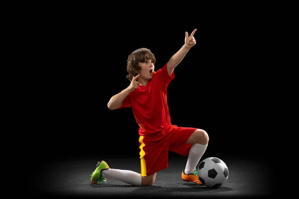 Winner. One little male football soccer player, boy training with football ball isolated on dark studio background. Concept of sport, game, hobby