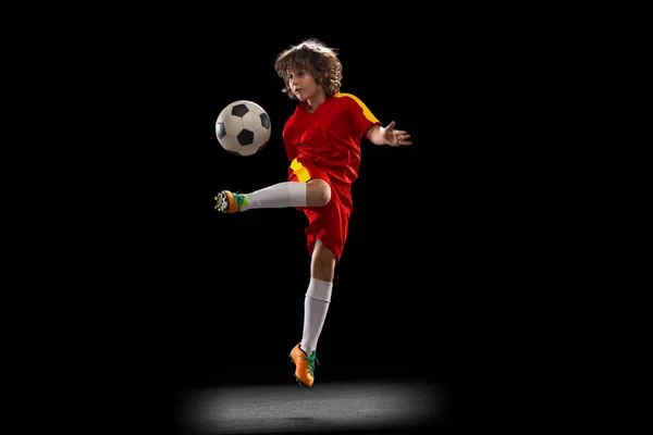 Один маленький футболіст, хлопчик тренується з футбольним м'ячем ізольовано на темному фоні студії. Концепція спорту, гри, хобі — стокове фото