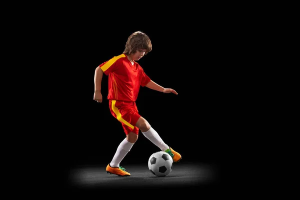 В движении. Маленький футболист, мальчик, тренирующийся с футбольным мячом, изолированный на тёмном фоне студии. Концепция спорта, игры, хобби — стоковое фото