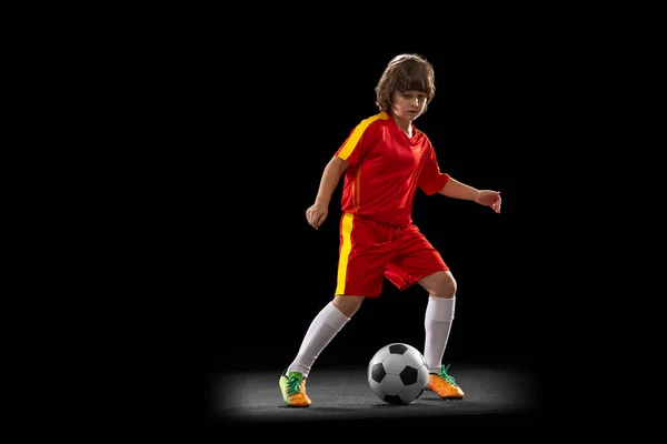 Menino pré-escolar, jogador de futebol em uniforme esportivo vermelho praticando com bola de futebol isolada no fundo escuro estúdio. Conceito de esporte, jogo, hobby — Fotografia de Stock