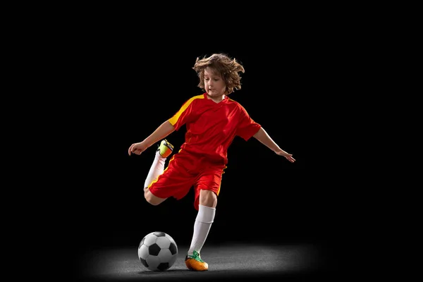 One little male football soccer player, boy training with football ball isolated on dark studio background. Concept of sport, game, hobby — Foto de Stock