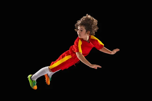 Preschool boy, football soccer player in red sports uniform practicing with football ball isolated on dark studio background. Concept of sport, game, hobby — Foto Stock