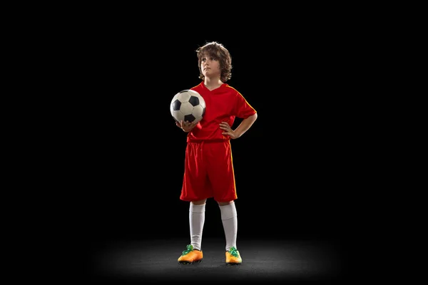 Portrait of little boy, football soccer player posing with football ball isolated on dark studio background. Concept of sport, game, hobby and childhood — Foto Stock