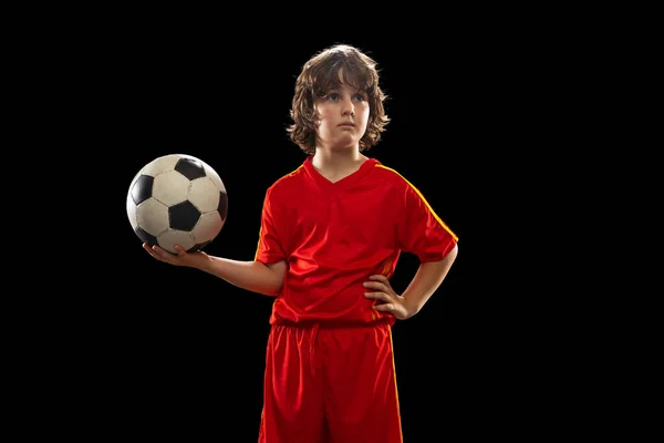 Retrato de menino, jogador de futebol posando com bola de futebol isolado no fundo escuro estúdio. Conceito de esporte, jogo, hobby e infância — Fotografia de Stock