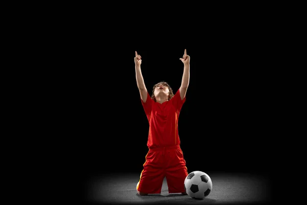 Vencedor. Um pequeno jogador de futebol masculino, treinamento de meninos com bola de futebol isolada no fundo do estúdio escuro. Conceito de esporte, jogo, hobby — Fotografia de Stock
