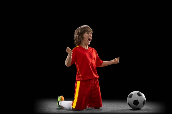 Happy, excited boy, football soccer player in red sports uniform with football ball isolated on dark studio background. Concept of sport, game, hobby — Foto Stock