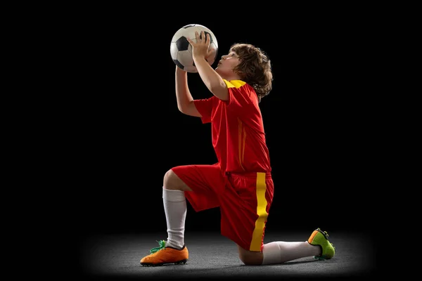 Happy, menino animado, jogador de futebol em uniforme de esportes vermelho com bola de futebol isolado no fundo escuro estúdio. Conceito de esporte, jogo, hobby — Fotografia de Stock