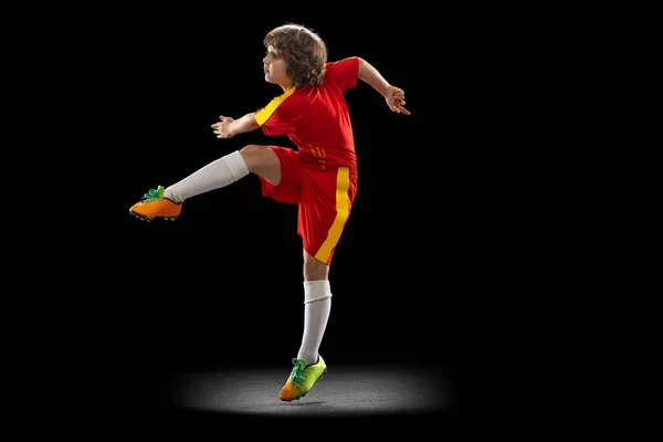 Preschool boy, football soccer player in red sports uniform practicing with football ball isolated on dark studio background. Concept of sport, game, hobby — Foto Stock