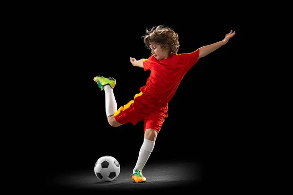 Em movimento. Um pequeno jogador de futebol masculino, treinamento de meninos com bola de futebol isolada no fundo do estúdio escuro. Conceito de esporte, jogo, hobby — Fotografia de Stock