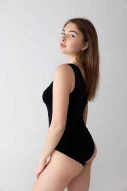 Gri stüdyo arka planında tek başına duran, siyah iç çamaşırlı, narin, sevimli bir kız. Doğal güzellik konsepti. Arka plan