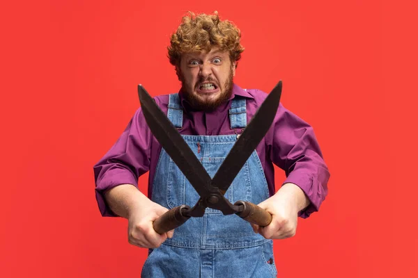 Close-up funny redheaded bearded man, farmer with big garden scissors isolated over red studio background. Concept of professional occupation, work. — Foto de Stock