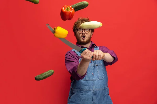Flying vegetables. Cute redheaded bearded man, farmer with knife having fun isolated over red studio background. Concept of professional occupation, work. — Stock Photo, Image